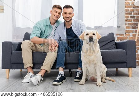 Same Sex Couple Sitting On Sofa Near Dog In Living Room.