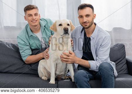 Cheerful Same Sex Couple Sitting On Couch With Dog.