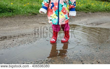 A Little Girl In A Bright Jacket And Red Boots Jumps Merrily Through Puddles. Spring Mood.