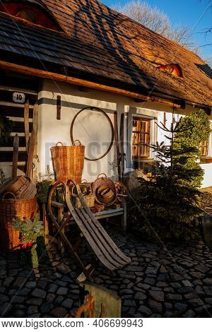 Old Traditional Sledge In Front Of Old House, Christmas Tree, Wicker Baskets On The Bench, Wooden Ro