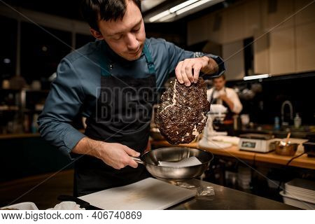 Male Chef In Black Apron Gently Holds Piece Of Cooked Meat And Frying Pan