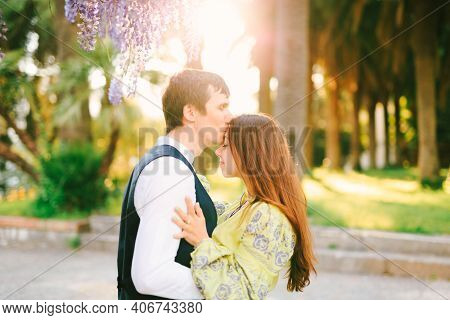 Man Is Kissing A Woman On The Forehead Under A Blooming Wysteria Tree In The Park