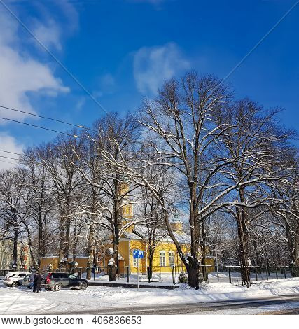 Latvia, Riga, February, 08, 2021 - Old Yellow Wooden Church Surrounded By Tall Trees On A Sunny Wint