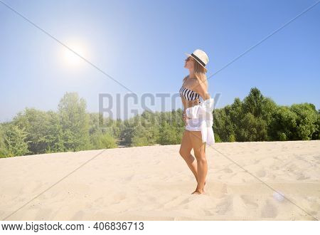 A View Of A Girl In A Top And A Hat Is Standing On The Sand Near The Forest. The Sun Shines Brightly