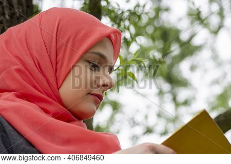 Cute Muslim Student Reading A Book While Sitting On The Tree Branch In The Park. Modern And Fashiona