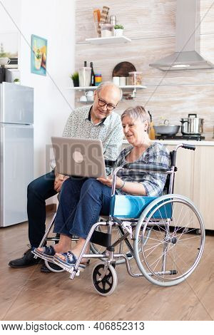 Paralyzed Senior Woman In Wheelchair And Husband Browsing On Internet Using Laptop In Kitchen. Disab