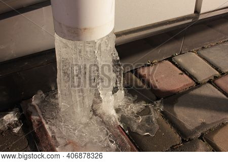 Frozen Water In A White Plastic Rain Pipe. Broken Icicles On Paving Slabs Background.