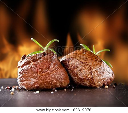 Pieces of red meat steaks with rosemary served on black stone surface. Blur fire flames on background