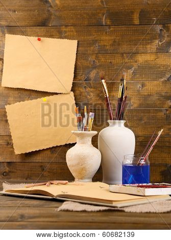 Composition with colorful watercolors, brushes in vase and sketcher on wooden background 