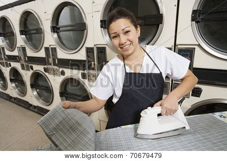 Portrait of a happy woman wearing apron ironing in front of washing machines