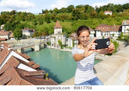 Travel selfie smart phone by woman in Bern Switzerland, Europe. Happy smiling multiracial Asian girl taking self portrait photo sitting on Nydegg bridge by the Aare river in the Swiss city of Berne