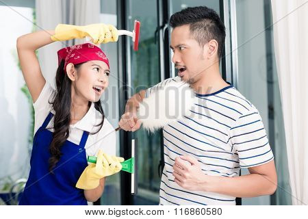 Asian woman and man having fun cleaning home staging a mock fight with chores utensils