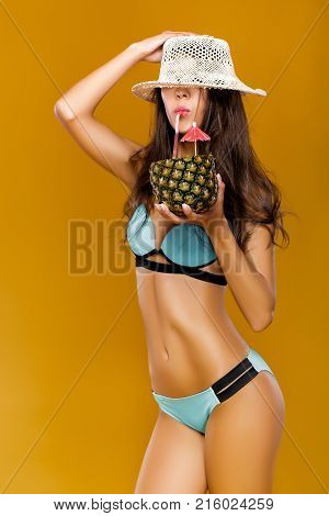portrait of beautiful woman in blue bikini drinks summer cocktail in pineapple on yellow background