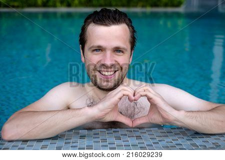 Handsome smiling young caucasian man makes the heart using fingers, hands during swimming at pool. Positive human emotions, feelings, body language. Concept of healthy lifestule and sport