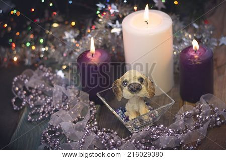 A romantic festive still life with burning white and purple candles and a toy puppy in a bowl with beads. A had crafted garland with a ribbon and beads. Purple colors. Dark Christmas background. Blurred bokeh