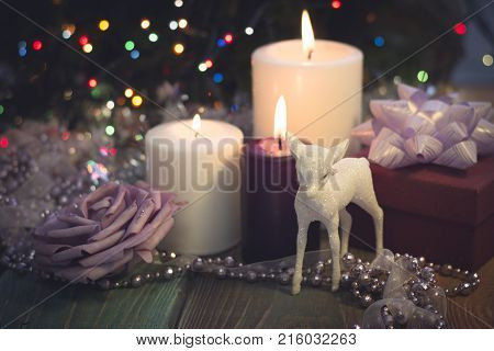 A festive still life with a purple and two white candles of different size, a burgundy gift box with a lilac bow, a light lilac garland of a ribbon and beads, a paper rose and a toy baby derr in a close up. Dark Christmas background. Blurred bokeh