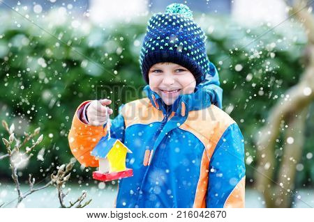 Little kid boy feeding birds in winter. Cute happy preschool child hanging colorful selfmade bird house on tree on frosty cold day. Preschooler in colorful wam clothes. Nature, empathy with animals.