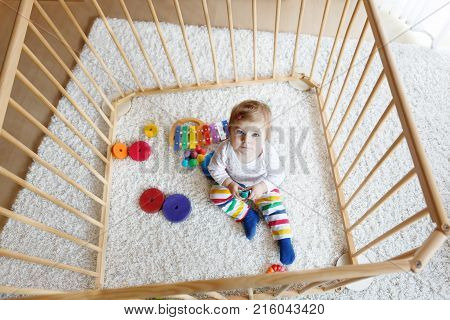 Beautiful little baby girl sitting inside playpen. Cute adorable child playing with colorful toys. Home or nursery, safety for kids.