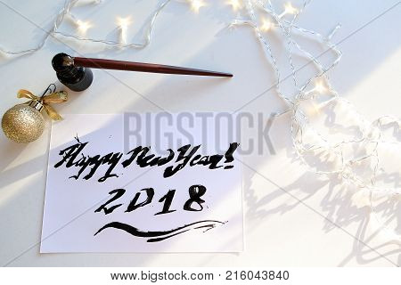 Christmas card with festive inscription Happy New Year, painted with black calligraphic font using carcass and fountain pen, which lies on table with Christmas garland in art workshop. Concept of beautiful writing, calligraphy and lettering, materials and
