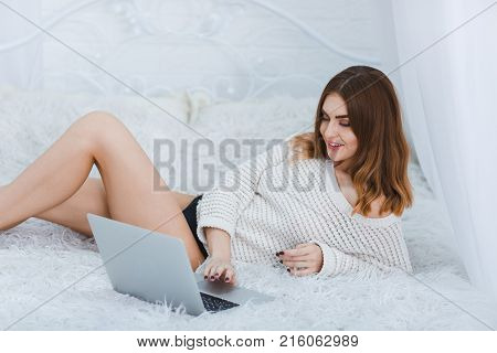 A young, beautiful girl in a white jacket lies on a bed leaning on her elbow with a laptop on a white background