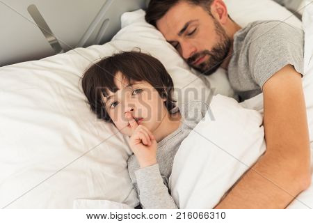 The boy asks not to wake his father. He put his forefinger to his lips and looks into the camera. His father had already fallen asleep. They lie alone in a clean white bed in their house.