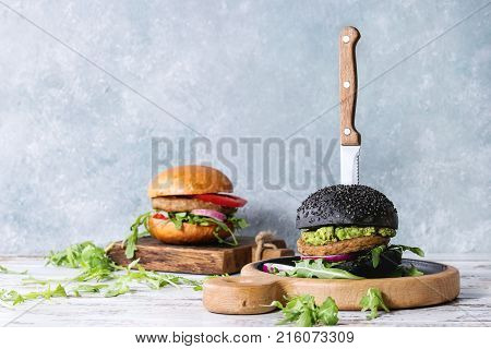Set of homemade burgers in black and white buns with avocado, tomato sauce, arugula, cheese, onion on wood serving board over white wooden plank table. Rustic style. Homemade fast food.