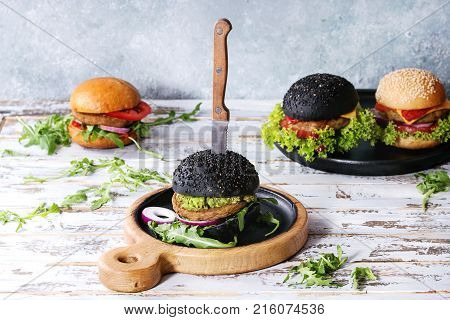 Set of homemade burgers in black and white buns with avocado, tomato sauce, lettuce, arugula, cheese, onion on wood serving board over white wooden plank table. Rustic style. Homemade fast food.