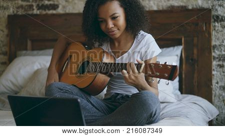 Attractive african american teenager girl concentraing learning to play acoustic guitar using laptop computer sitting on bed at home