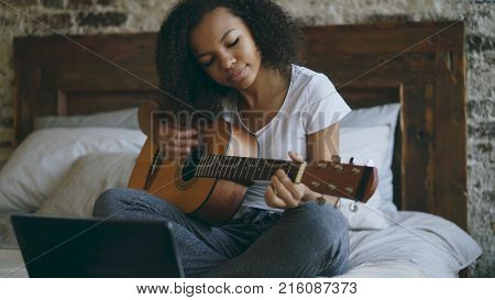 Attractive african american teenager girl concentraing learning to play acoustic guitar using laptop computer sitting on bed at home