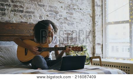 Curly african american teenager girl concentraing learning to play acoustic guitar using laptop computer sitting on bed at home