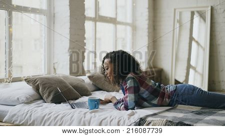 Curly mixed race student girl lauging while using laptop for sharing social media lying in bed at home