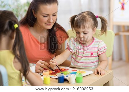 Children painting in preschool. Teacher helps by kid little girl.