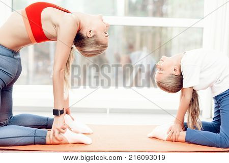 Mother and daughter are engaged in yoga in sportswear. They are in a bright room with panoramic windows. The in a ushtrasana asana, camel pose