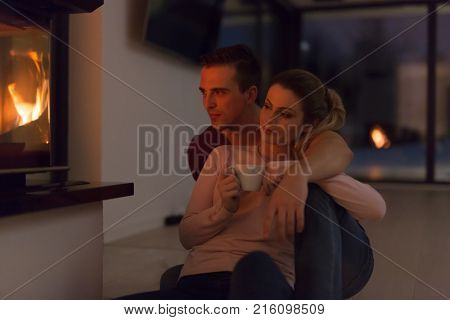 Young romantic couple sitting on the floor in front of fireplace,talking and drinking tea on cold winter night at home