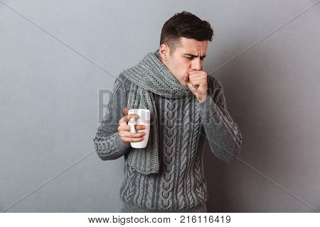 Sick Man in sweater and scarf holding cup of tea while having cough over gray background