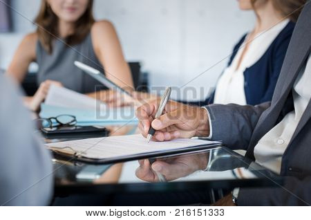 Closeup of hand of businessman holding pen and analyzing report during board meeting. Business people writing on document during annual meeting. Detail hand of business man fill the business contract.