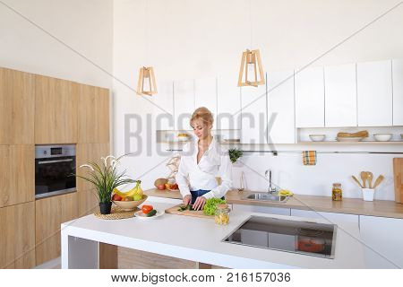 Attractive young woman cuts green cucumber for salad with sharp knife, takes red juicy tomato and several times throws up with smile on face, sniffs freshness of lettuce leaves and takes up shredding vegetables. Blonde at top of modern light kitchen with 