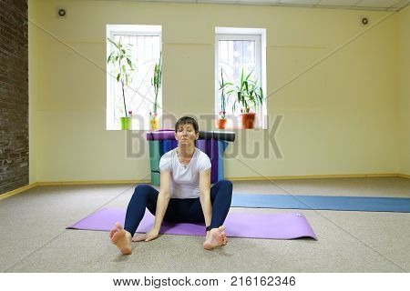 Sportswoman focused on stretch before Pilates class. Girl doing warm up for legs on mat. Female has hair pulled back, concentrated look. Athlete dressed in dark top, yoga legging trains in fitness room. Concept of online training, customizable workouts.
