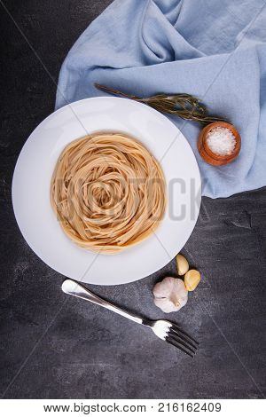 Spaghetti of hard varieties in a white plate, on dark concrete, next to fork, garlic and salt, the plate stands on a blue fabric. View from above.
