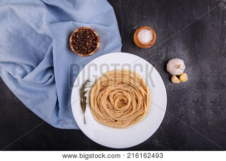 Spaghetti of hard varieties in a white plate, with a sprig of marble, on dark concrete, next to it lies garlic, salt, and picking peppers in peas, a plate stands on a blue fabric. View from above.