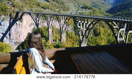 Brunette girl with long hair sitting at the table and enjoying the view of the bridge Durdevica Tara bridge in Montenegro. A break during long trips. Focus on the bridge