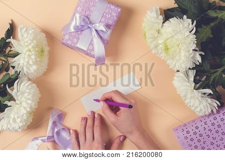 woman hand writing a note with the text thank you on a paper, over beige table and flowers. toned