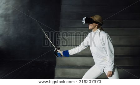 Concentrated young fencer woman practice fencing exercises using VR headset and training simulator competition game indoors