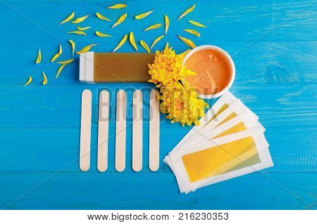 Set for depilation from strips for depilation, wax in dishes and wooden sticks. Yellow chrysanthemums on a blue wooden background. View from above