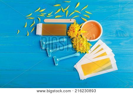 Set for depilation with strips, chopsticks, wax in a cup and wax in a cassette and a row of razors with chrysanthemums on a blue wooden background. View from above