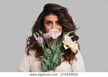 Beautiful as a flower. Attractive young woman covering face with bunch of flowers and looking at camera while standing against grey background