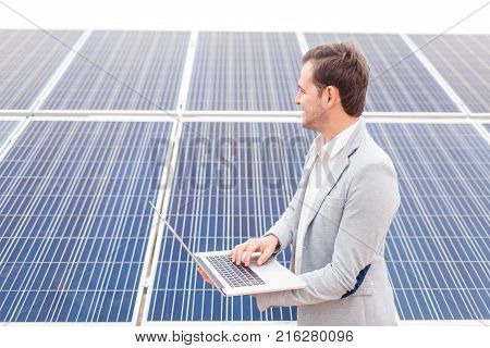 A happy man in a suit holds an open modern laptop in his hands and looks in the distance standing next to solar panels. Outdoors