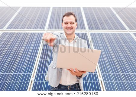 Happy handsome man in costume holding hands open modern laptop smiling and showing index finger in front of him, standing next to solar panels. Outdoors