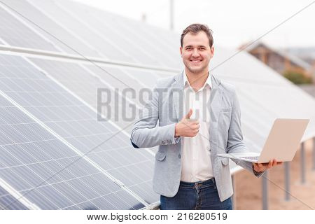A smiling man of European appearance in jeans and a jacket stands on a background of solar panels, holds in his hands an open modern laptop and shows a thumbs up. Outdoors