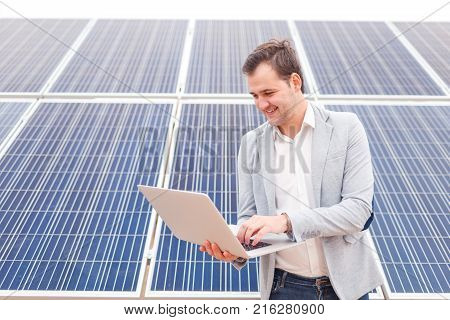 Smiling man working in a jacket holds in his hands an open modern laptop standing next to solar panels. Outdoors.
